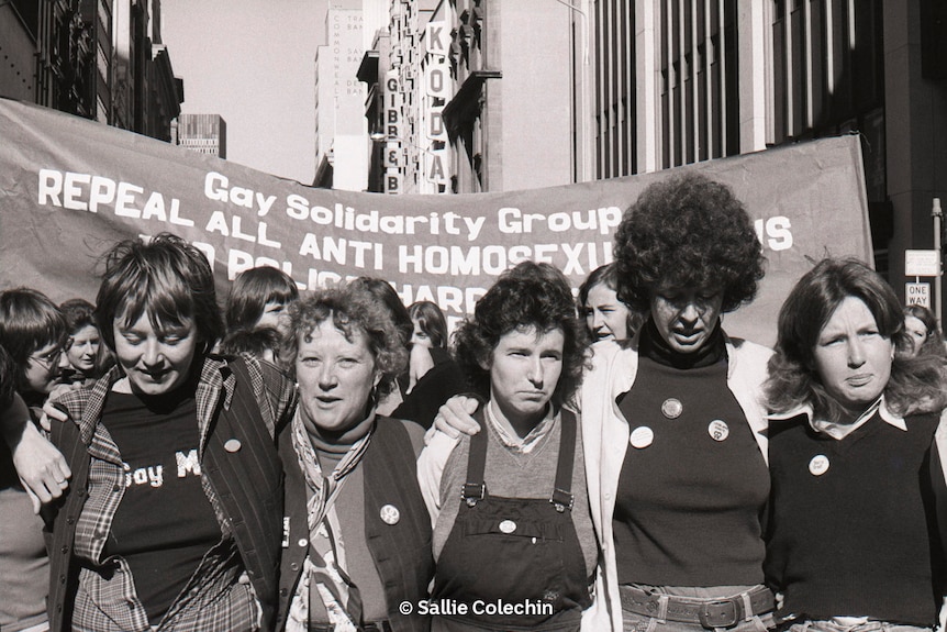 A black-and-white image of four women, standing shoulder-to-shoulder, if front of a banner reading "gay solidarity group"