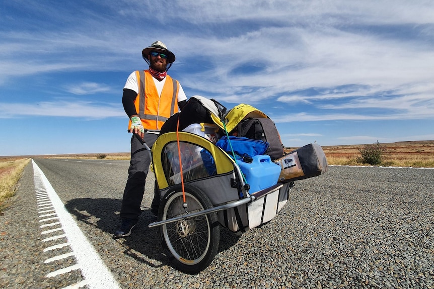 A mean wearing a high vis vest and fly net hat pushes a heavily-laden hand cart along a long straight road.