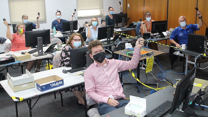 Nine people sitting at desks in front of computers holding up phones, all wearing masks.