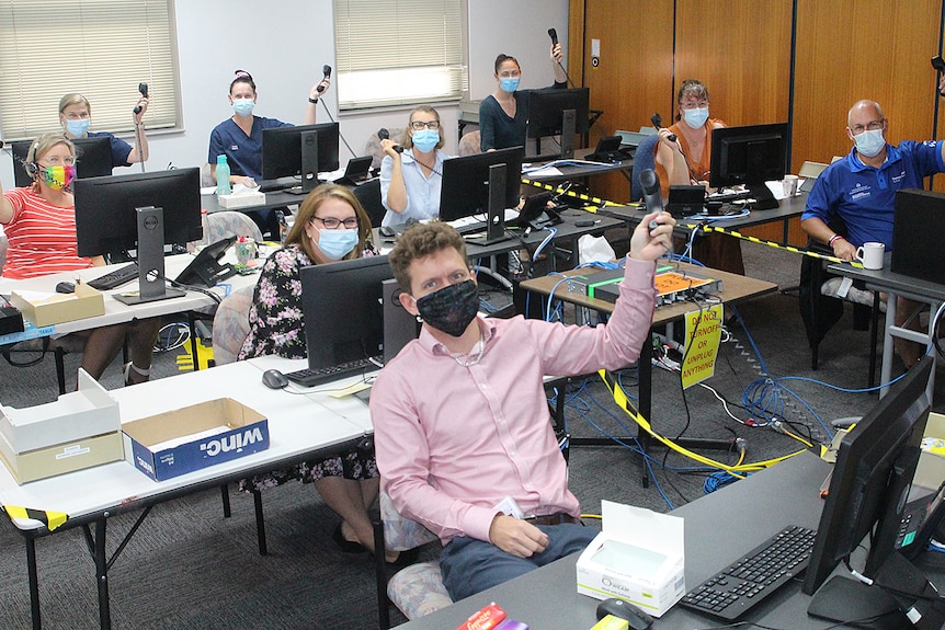 Nine people sitting at desks in front of computers holding up phones, all wearing masks.