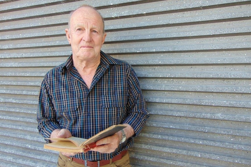 Man with balding grey hair wearing checked shirt and brown jeans and holding a small book.