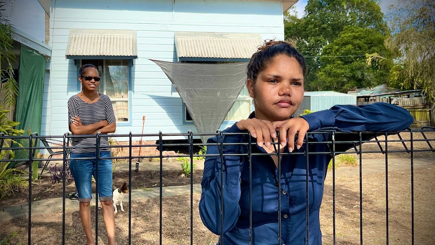 A young woman leans on a fence while another stands in the front yard of a house behind her.