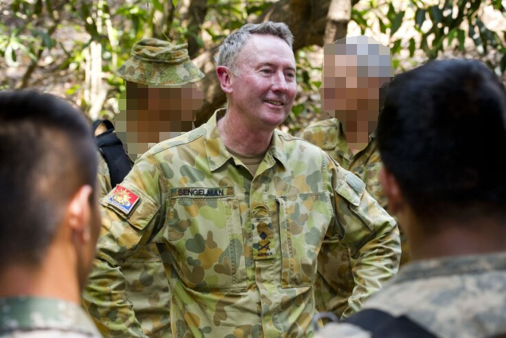 A man smiling, standing in military fatigues with other soldiers in front and behind of him in a forest location.