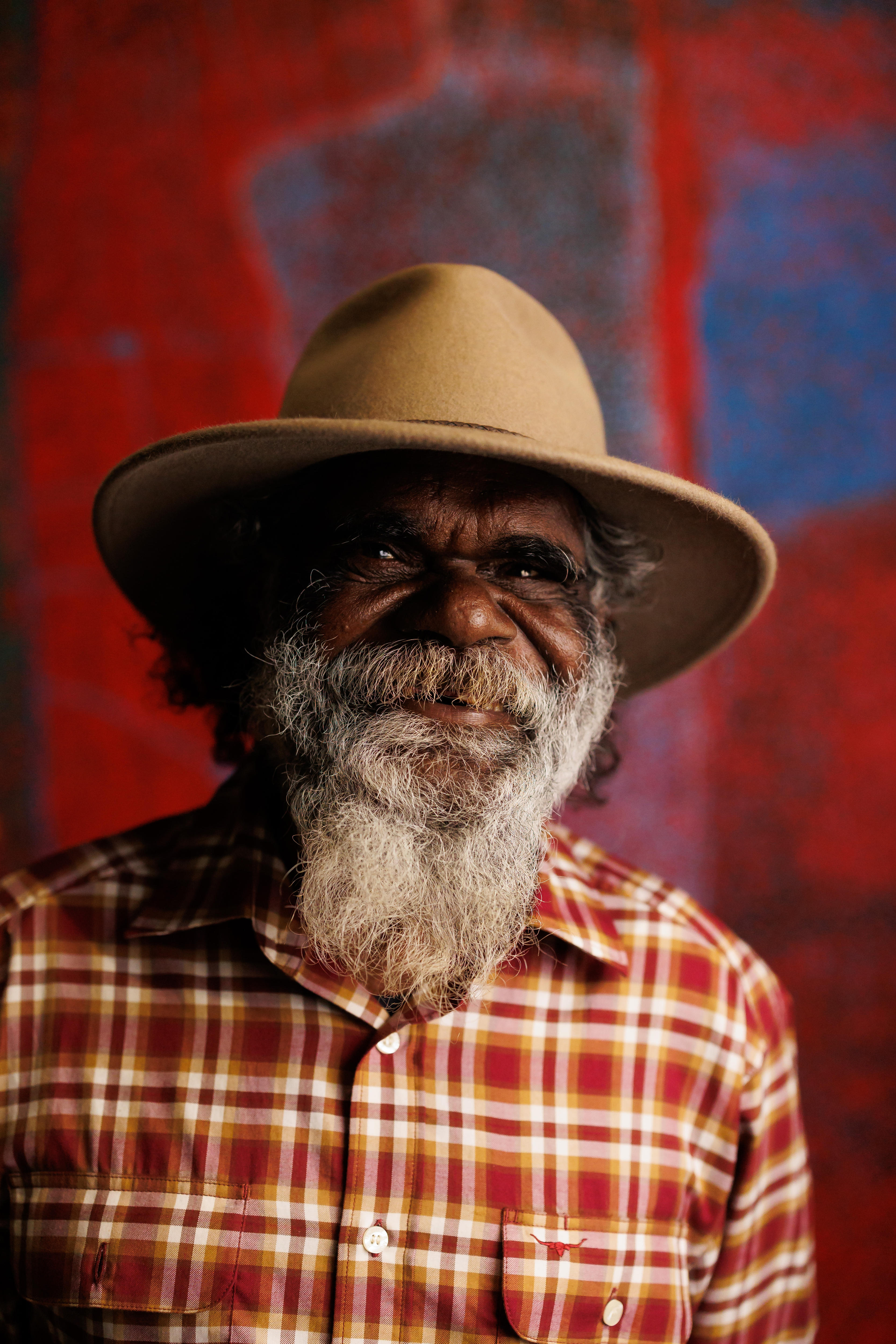 Noli Rictor, an Indigenous Elder with a white beard, wearing a hat, stands smiling in front of a red painting.