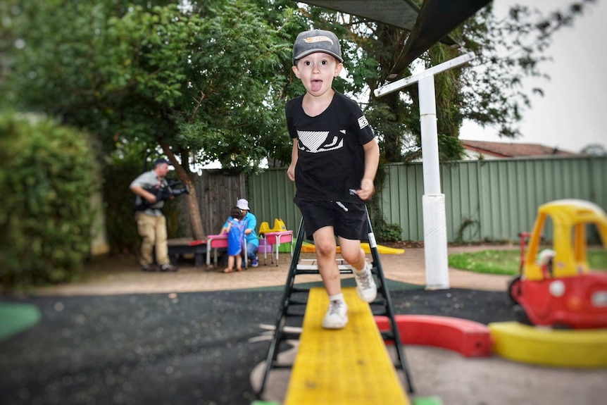 Young boy with big smile running along yellow plank at day care, colourful toys in background.