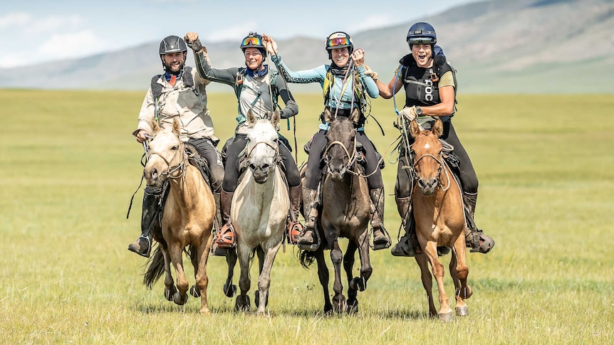 Four horse riders gallop across a green paddock under a blue sky holding hands and celebrating.