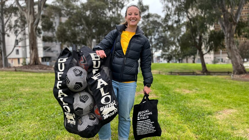 A woman holding soccer balls and smiling in yellow shirt and black puffy jacket. 