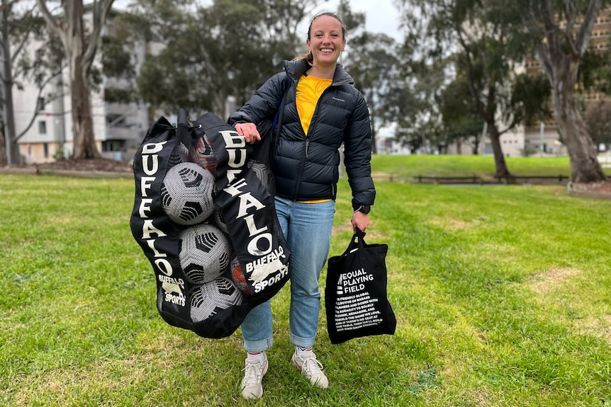 A woman holding soccer balls and smiling in yellow shirt and black puffy jacket. 