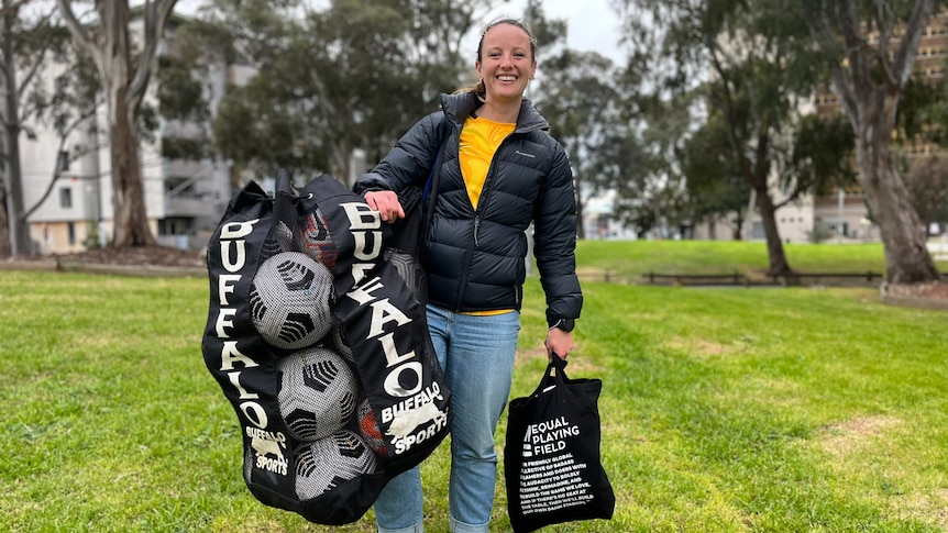 A woman holding soccer balls and smiling in yellow shirt and black puffy jacket. 