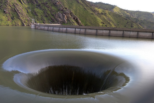 A giant drain sucking water out of a lake with a dam wall and green hills behind it