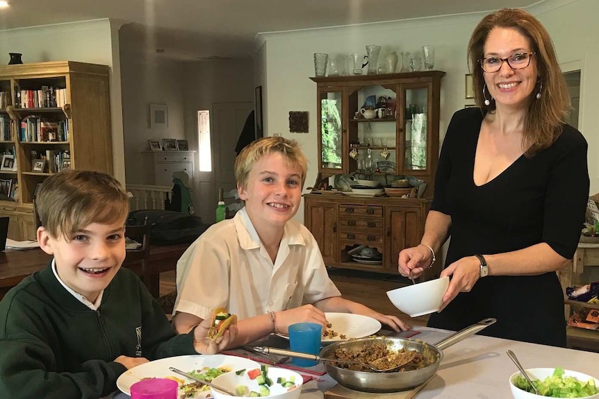 A mother stands smiling at her kitchen as she makes dinner. Her two young boys sit at the table eating.