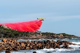An aerial water bomber drops fire retardant on bushland with the ocean and rocks in the foreground.