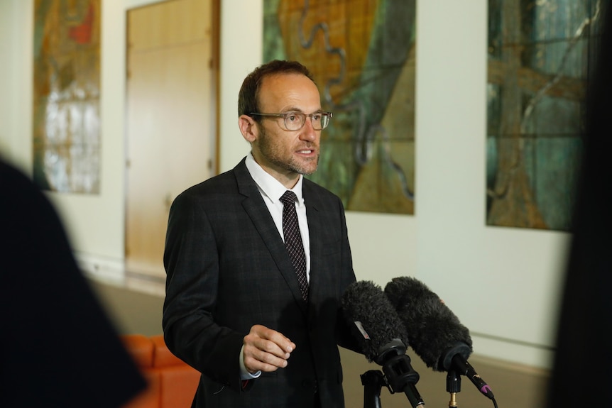 Politician Adam Bandt stands inside a building, speaking into two microphones while wearing a suit and glasses