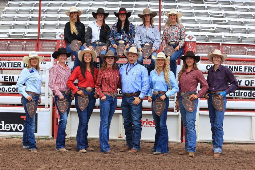 A man and 12 women, all dressed in cowboy outfits, pose for a photo in a rodeo stadium.