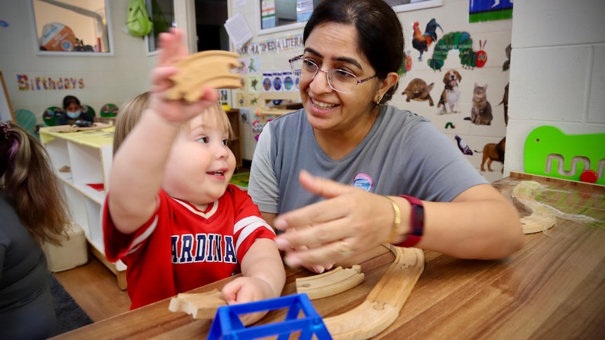 A female childcare worker sitting next a young toddler who is playing with toys