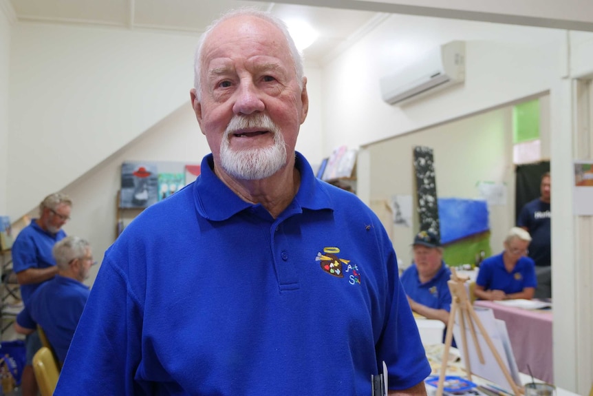 A man with grey hair stands smiling, in front of people doing artwork.