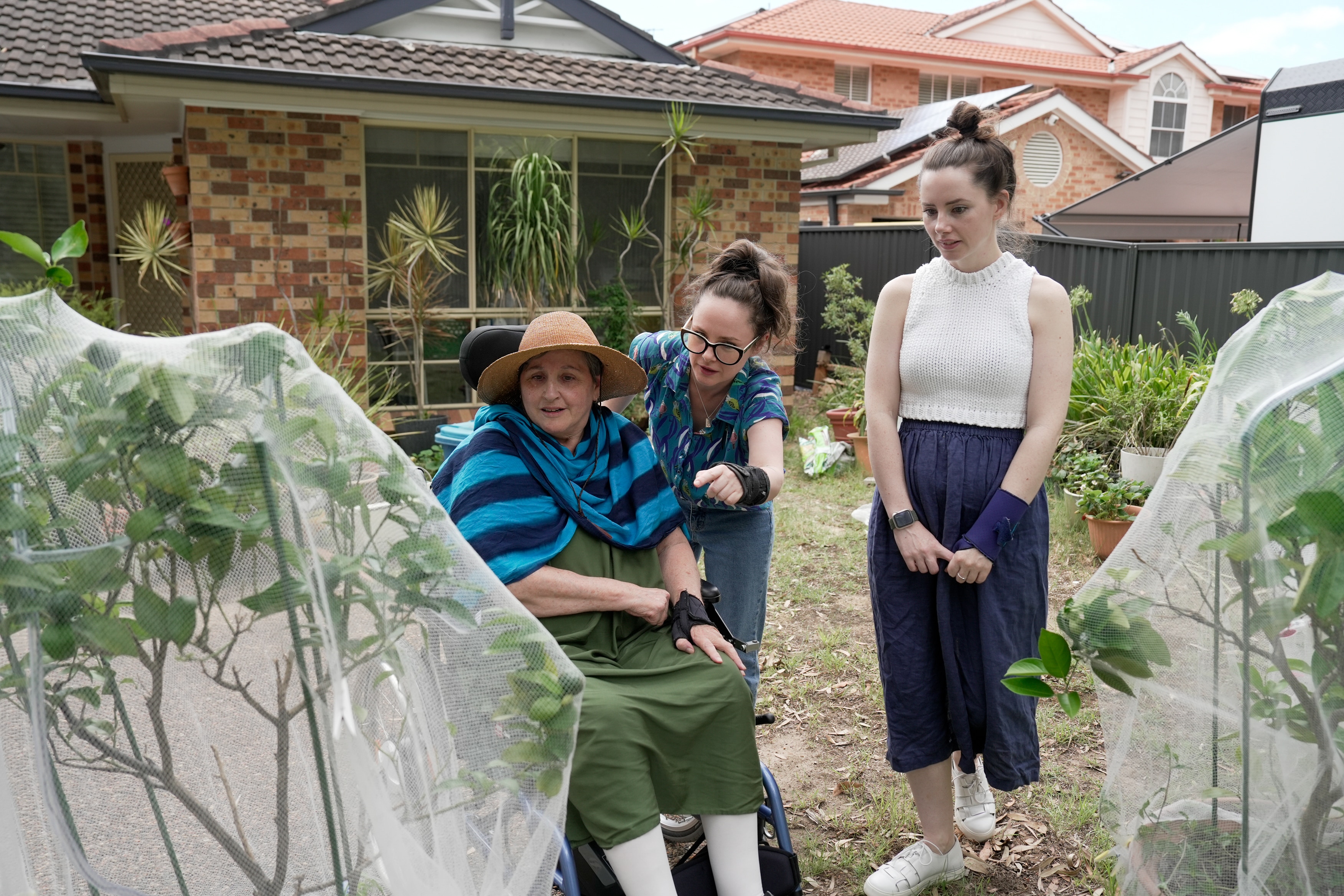 Una mujer blanca de mediana edad sentada en una silla de ruedas en un jardín, flanqueada por sus dos hijas adultas.