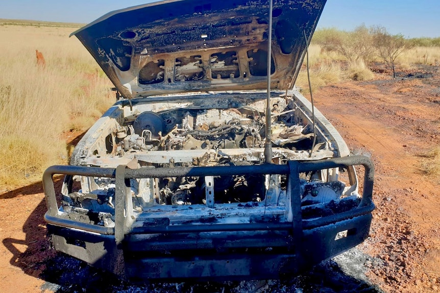 The bonnet of a burnt-out car wreck sits open on a gravel track
