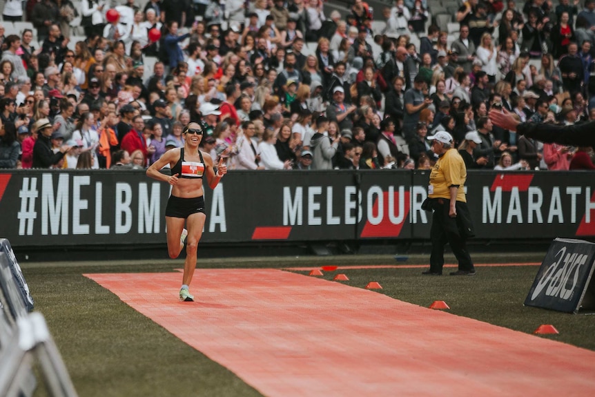 A smiling female runner gives a thumbs up on a running track with a big crowd behind her.