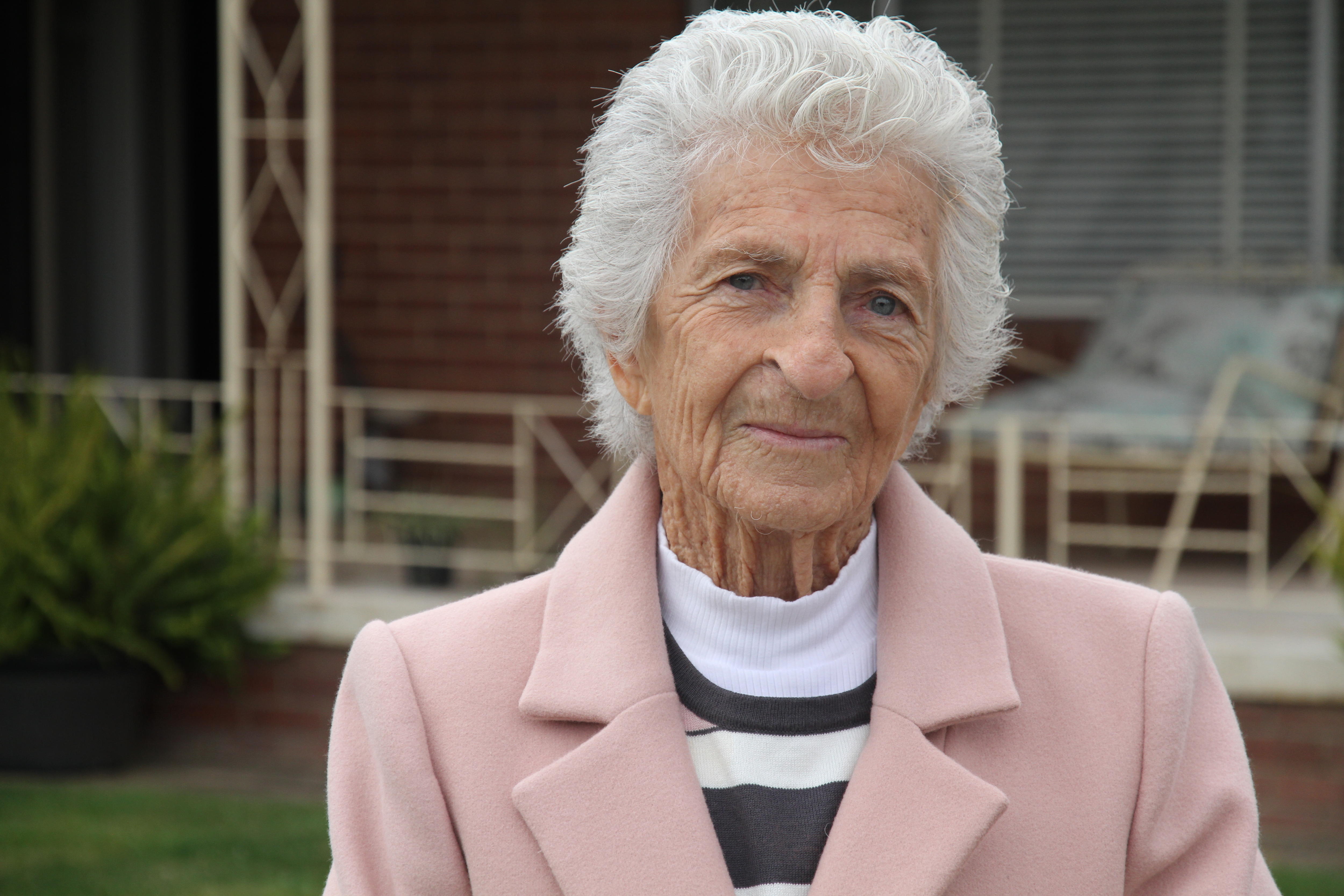 A serious elderly woman with short grey hair, whtie and grey top, and pink coat in front of old veranda.