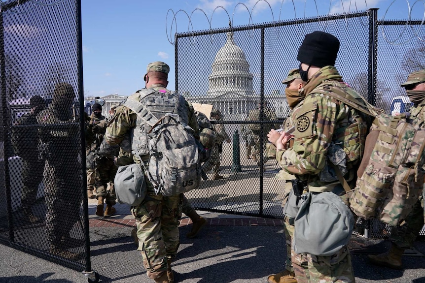 National Guard troops walk between a black security fence with the US Capitol building in the background