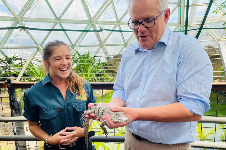 Scott Morrison holds a small crocodile in two hands while a female zookeeper laughs while standing next to him.
