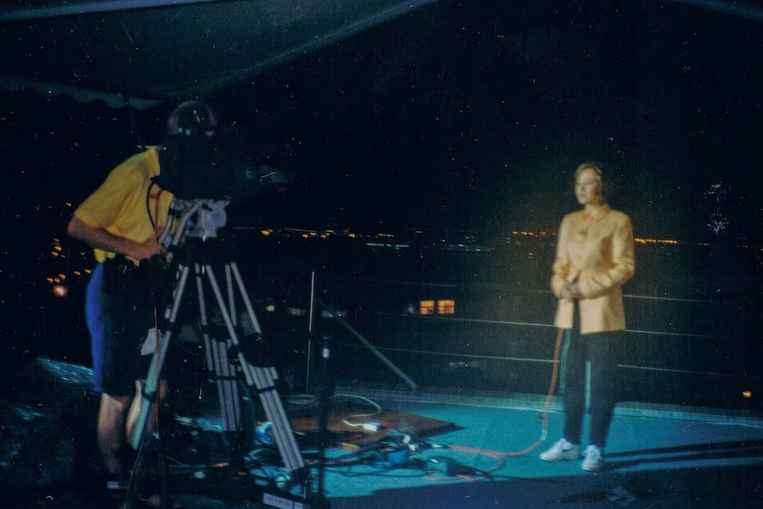 A woman reporter stands in front of a television camera in the dark, overlooking Hong Kong.