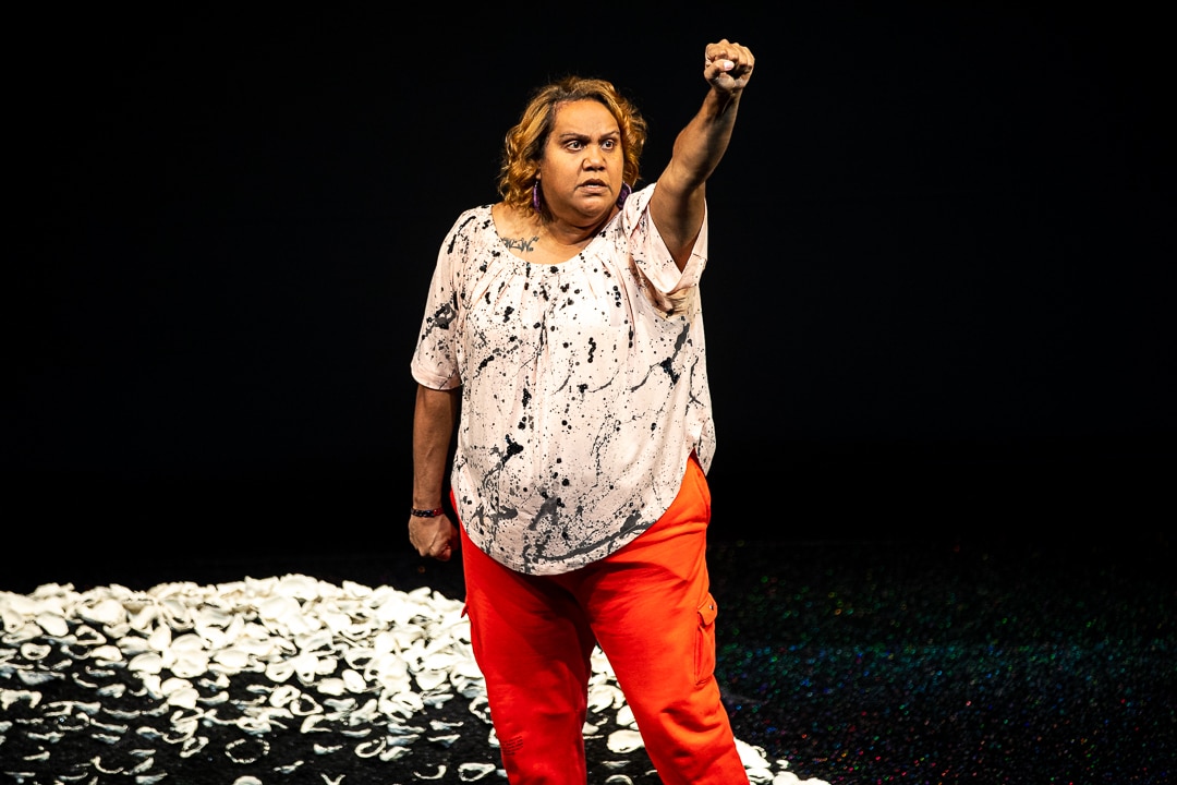 Elaine Crombie with her fist raised in the air in front of midden shells on mound of dirt in The 7 Stages of Grieivng