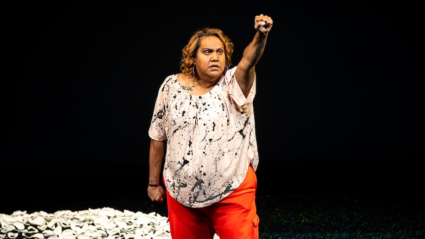 Elaine Crombie with her fist raised in the air in front of midden shells on mound of dirt in The 7 Stages of Grieivng