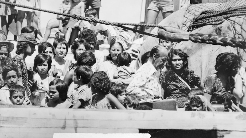 Black and white photo of large crowd of people, many squinting into the sun, aboard a boat, just visible.