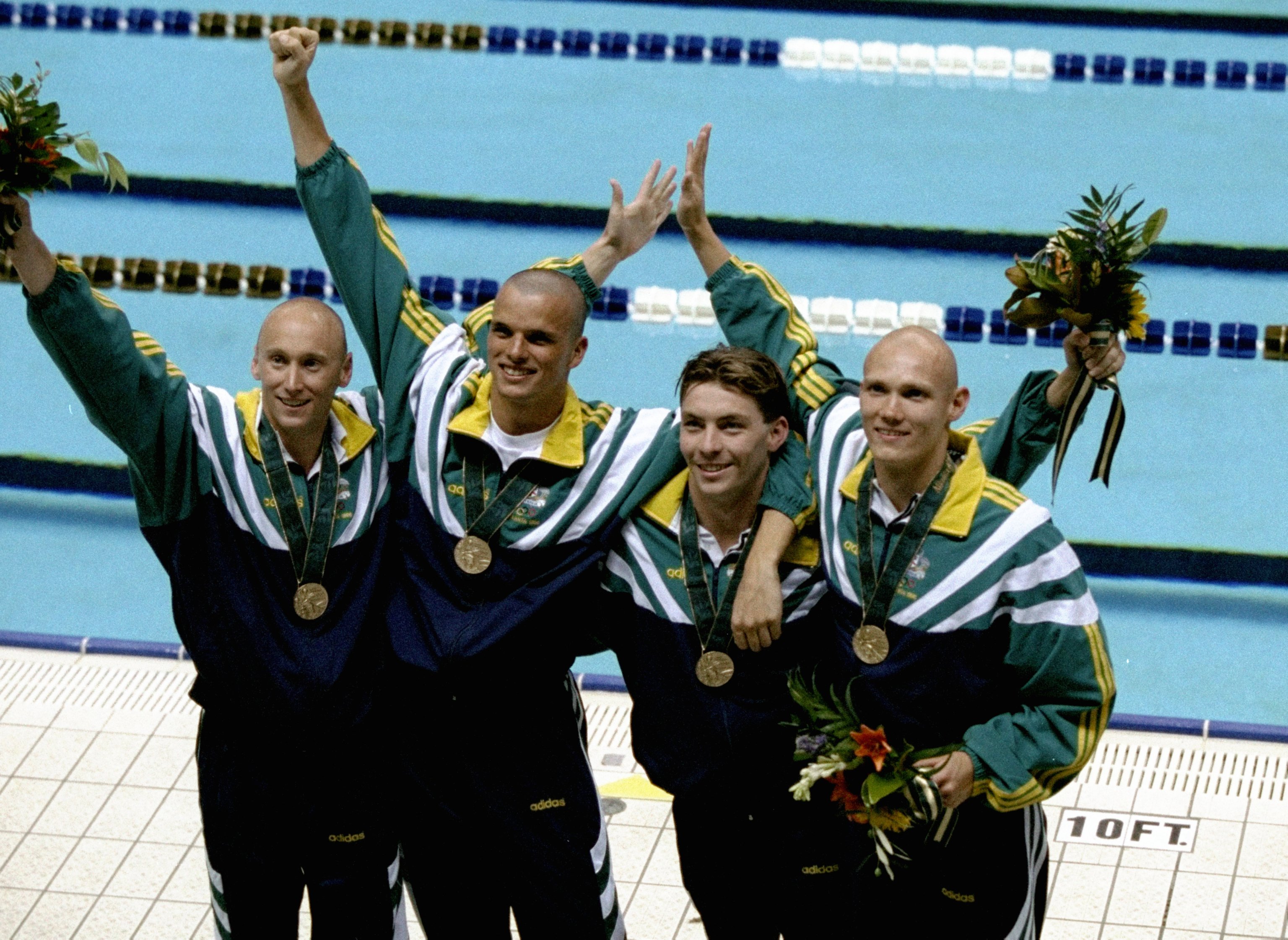 Four men wearing green and gold tracksuits stand in front of a pool with bronze medals around their neck.