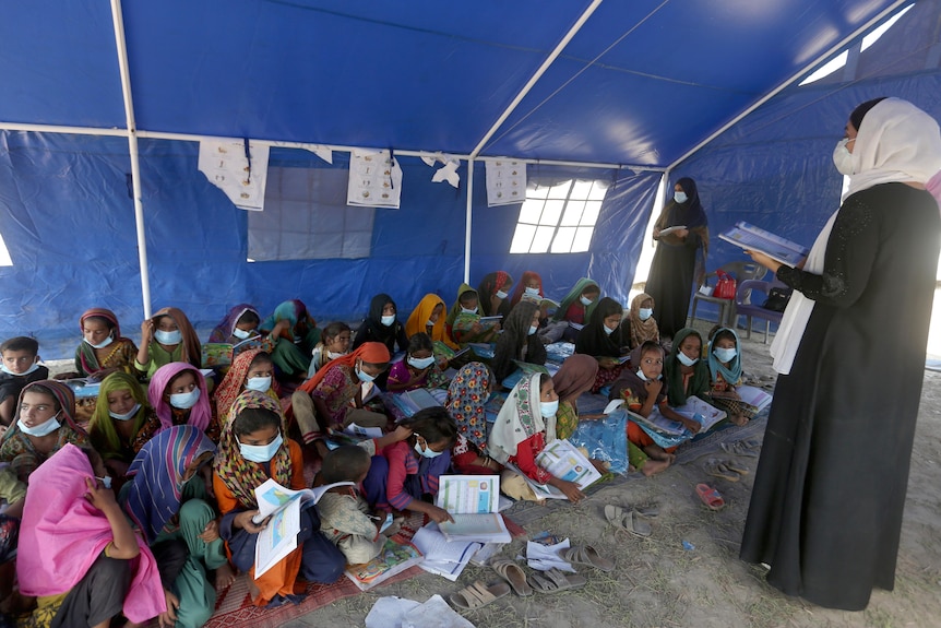 A group of schoolchildren sit packed together on the ground with textbooks in a blue tent, listening to a teacher.
