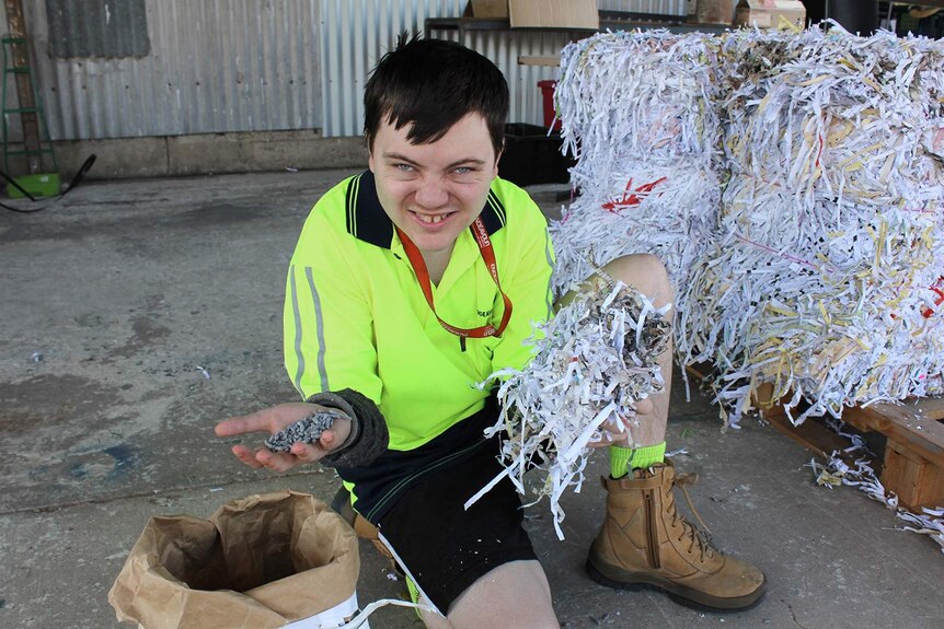A young man in a high visibility shirt holds up handfuls of shredded paper and kitty litter pellets made from paper