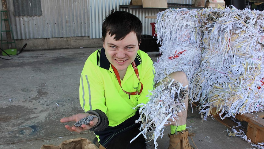 A young man in a high visibility shirt holds up handfuls of shredded paper and kitty litter pellets made from paper