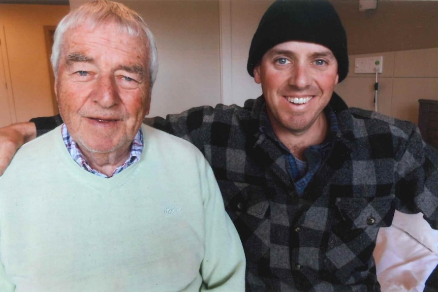 An old man in an aqua blazer smiles next to a younger man wearing a beanie and flannel shirt, smiling.