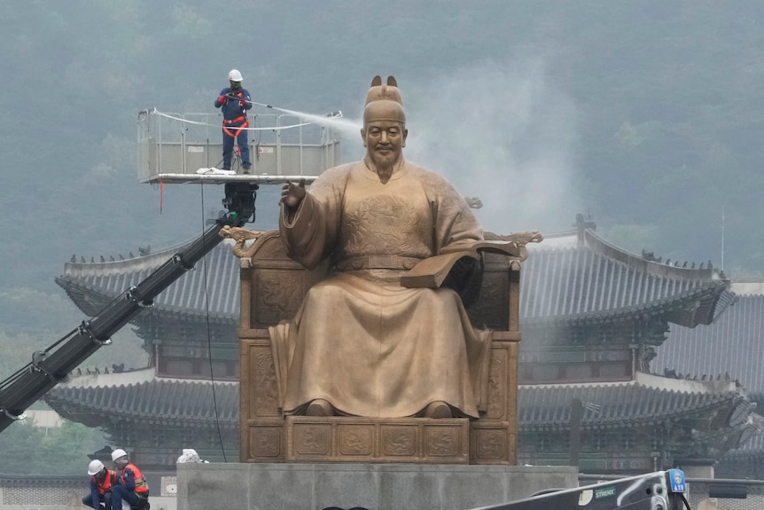 A worker on an elevated platfrom cleans a giant statue of an ancient Korean king with a pressure washer. 
