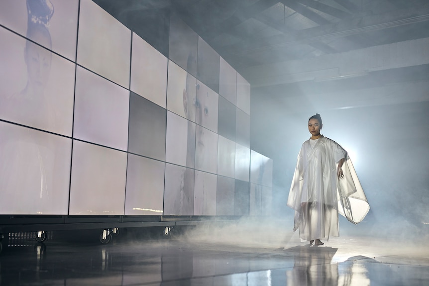 A dancer in all white walks through a plume of smoke beside a checkered panel of projection screens.