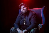 Colour portrait of rapper Briggs sitting on a chair in front of black background and some red light.