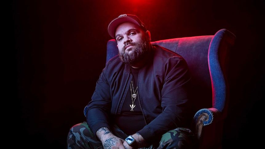 Colour portrait of rapper Briggs sitting on a chair in front of black background and some red light.