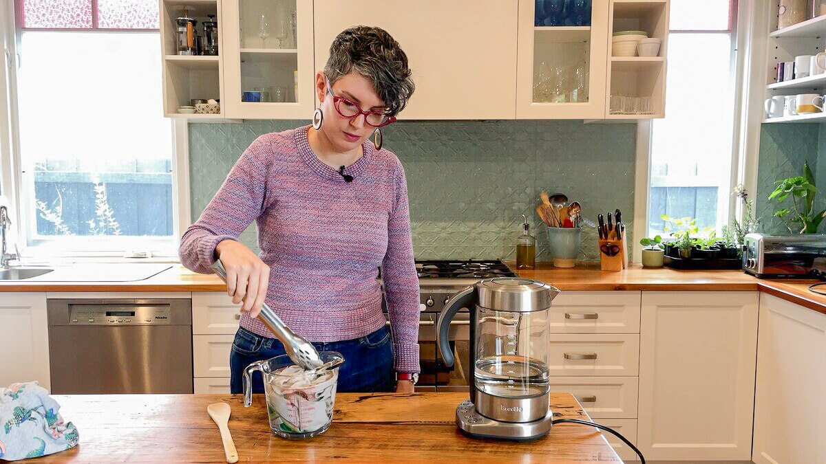 Woman in her kitchen in front of wooden bench holding tongs, checking on soaking stained garment in bowl of boiling water.