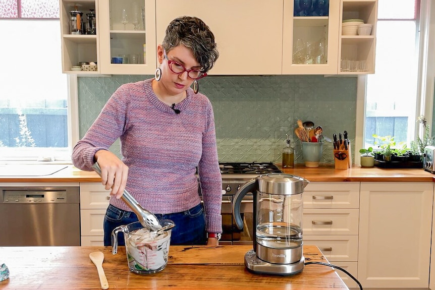 Woman in her kitchen in front of wooden bench holding tongs, checking on soaking stained garment in bowl of boiling water.