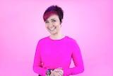 A woman with short pink and purple hair smiles to the camera. She wears a pink top and the background is pink