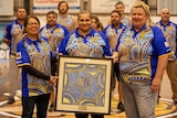 Three ladies with an Indigenous painting, with seven males in background, all wearing Indigenous uniform.