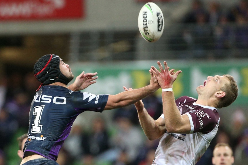 Two male NRL players jump in the air as they try to catch a football.