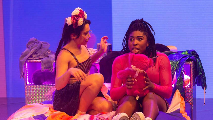 Two young woman in bright outfits sit excitedly on a bed on stage