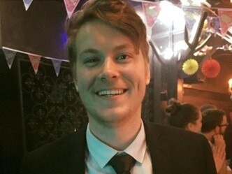 A profile shot of Dylan wearing a black suit, white shirt and black tie at an indoor party with bunting and decorations.