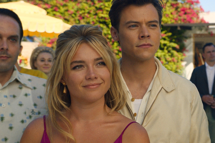 Blonde white woman with pink dress stands beside brunette white man in taupe jacket on a sunny neighbourhood street.