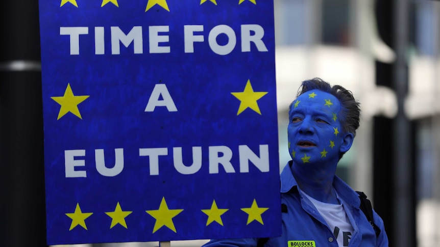 A man with his face painted blue with a circle of yellow stars holds a sign reading 'time for a EU turn'