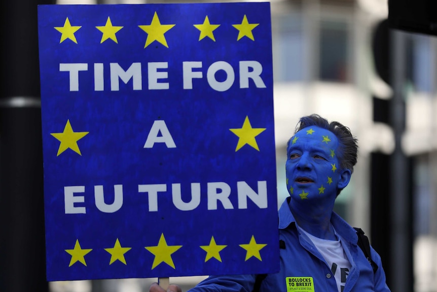 A man with his face painted blue with a circle of yellow stars holds a sign reading 'time for a EU turn'