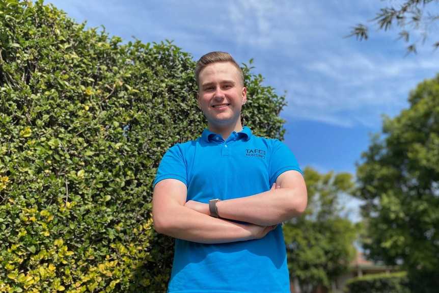 A young man in a blue shirt stands next to a hedge.